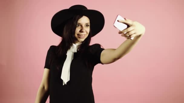 女性はスマートフォンで自分撮りを取ります。ピンクの背景に携帯電話と黒いドレスと帽子の女性. — ストック動画