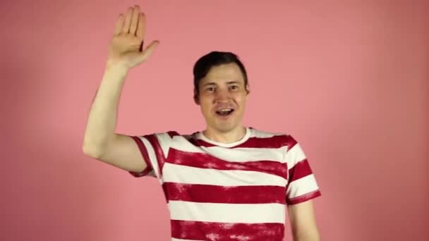 A man in a striped t-shirt on a pink background. Portrait of a cheerful young man, waving his hand — Stock Video