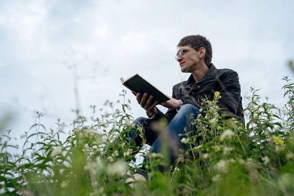 Vue latérale de jeune homme beau dans des lunettes et veste chaude livre de lecture dans l'espace ouvert sur fond de collines verdoyantes — Photo
