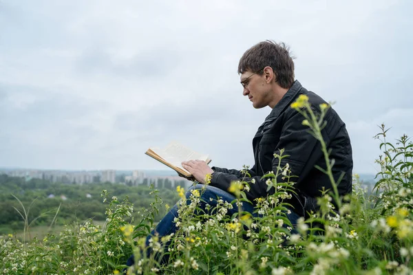 Zijaanzicht van jonge knappe man in glazen en warme jas lezen boek in open ruimte op de achtergrond van groene heuvels — Stockfoto