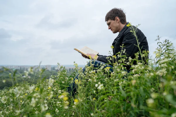 Zijaanzicht van jonge knappe man in glazen en warme jas lezen boek in open ruimte op de achtergrond van groene heuvels — Stockfoto