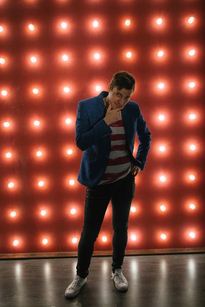 Mladý muž stojí u osvětlené stěny. Trendy muž dívá na kameru na červeném pozadí se žárovkami — Stock fotografie