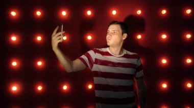 Genç bir erkek aydınlanmış bir duvarın önünde duruyor ve akıllı telefondan selfie çekiyor.