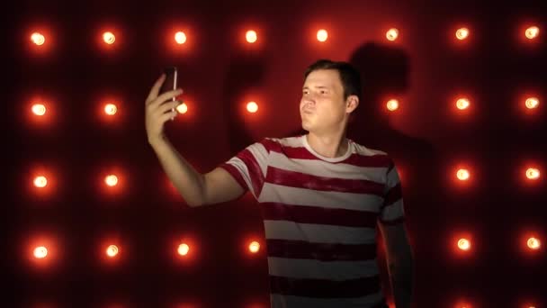 Молодий чоловік стоїть проти освітленої стіни і бере селфі на смартфон — стокове відео
