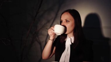 Elinde bir fincan kahve olan bir kadın. Genç hayalperest kadın, karanlıkta ağacın dallarının yanında ışığın altında dururken kahvenin tadını çıkarıyor.