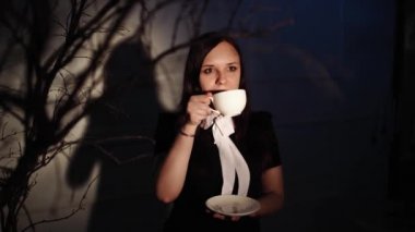 Elinde bir fincan kahve olan bir kadın. Genç hayalperest kadın, karanlıkta ağacın dallarının yanında ışığın altında dururken kahvenin tadını çıkarıyor.