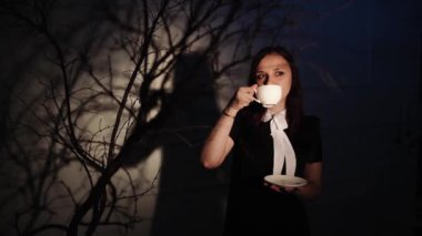 Elinde bir fincan kahve olan bir kadın. Genç hayalperest kadın, karanlıkta ağacın dallarının yanında ışığın altında dururken kahvenin tadını çıkarıyor.