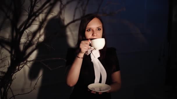 Mujer con taza de café en lugar oscuro. Joven mujer de ensueño disfrutando del café mientras está de pie en un rayo de luz junto a ramas de árbol en la oscuridad — Vídeos de Stock