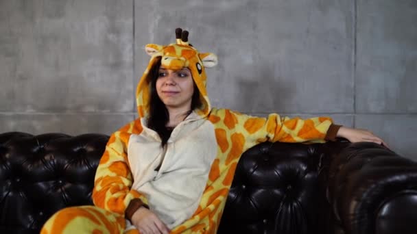 Zsiráfjelmezes nő ül a kanapén. Mosolygó fiatal nő vicces pizsamában zsiráf ül a bőr kanapén, és a kamerába néz — Stock videók