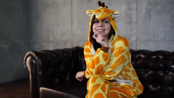Mulher vestida de girafa sentada no sofá. Sorrindo jovem mulher em pijama engraçado de girafa sentado no sofá de couro e olhando para a câmera — Vídeo de Stock