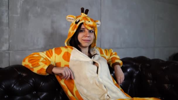 Женщина в костюме жирафа сидит на диване. Улыбающаяся молодая женщина в смешной пижаме жирафа сидит на кожаном диване и смотрит в камеру — стоковое видео