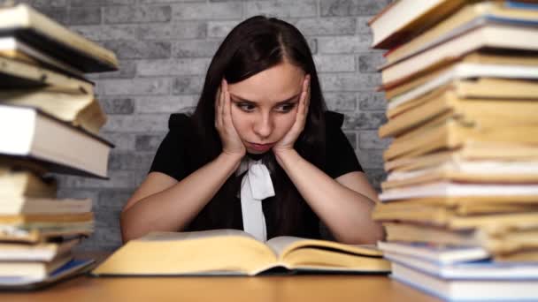 Vermoeide vrouwelijke student lezing tussen boeken. Pensive jonge vrouw zittend aan tafel met stapel boek en lezing op achtergrond van grijze muur — Stockvideo