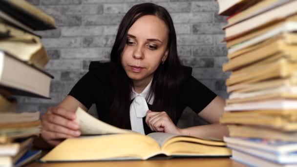 Estudiante cansada leyendo entre libros. Mujer joven pensativa sentada a la mesa con pila de libro y lectura sobre fondo de pared gris — Vídeo de stock