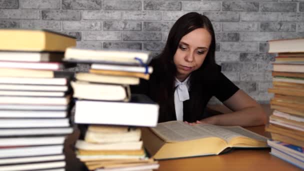Trött kvinnlig elev läsning bland böcker. Pensive ung kvinna sitter vid bordet med högen av bok och läsning på bakgrund av Grey Wall — Stockvideo