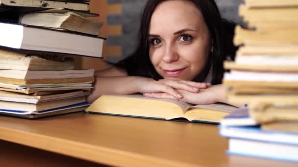Fáradt női hallgató olvasatban könyvek között. Pensive fiatal nő ül az asztalnál halom könyv és olvasás a háttérben a szürke fal — Stock videók