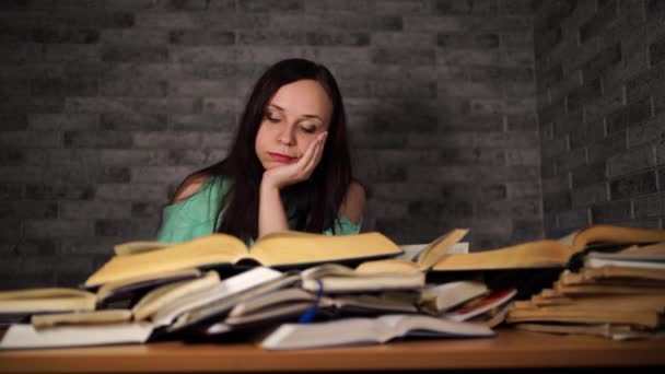 Fatiguée de lire parmi les livres. Jeune femme pensive assise à table avec pile de livre et lecture sur fond de mur gris — Video