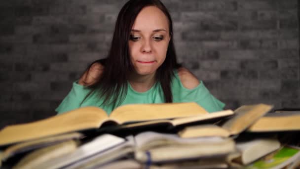 Zmęczona studentka czytania wśród książek. Pensive młoda kobieta siedzi przy stole z kupą książki i czytania na tle szarej ściany — Wideo stockowe