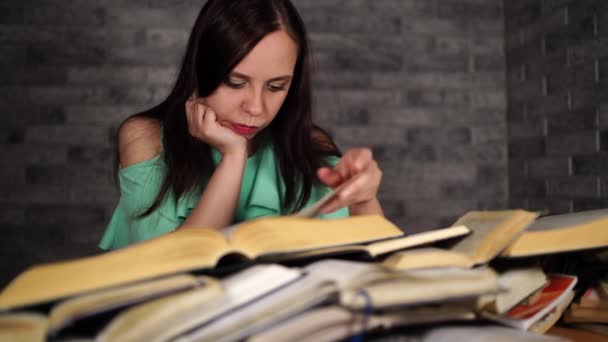 Estudiante cansada leyendo entre libros. Mujer joven pensativa sentada a la mesa con pila de libro y lectura sobre fondo de pared gris — Vídeo de stock