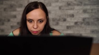 Young woman looking at display of laptop. Concentrated brunette in blouse with naked shoulders browsing while working on computer against grey wall