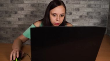 Young woman looking at display of laptop. Concentrated brunette in blouse with naked shoulders browsing while working on computer against grey wall