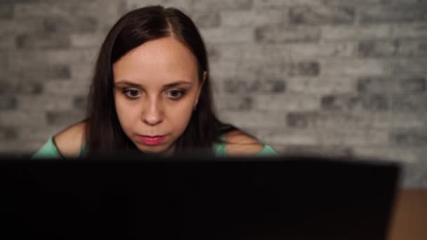 Jonge vrouw kijkt naar vertoning van laptop. Geconcentreerde brunette in blouse met naakte schouders browsen tijdens het werken op de computer tegen grijze muur — Stockvideo