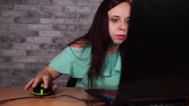 Молодая женщина смотрит на дисплей ноутбука. Сконцентрированная брюнетка в блузке с обнаженными плечами просматривает во время работы за компьютером против серой стены — стоковое видео