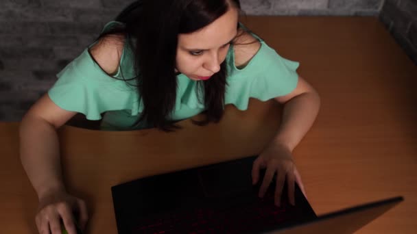 Junge Frau am Tisch sitzend und mit Laptop. Konzentrierte Brünette schaut auf die Anzeige des Laptops, während sie auf dem Hintergrund einer grauen Wand arbeitet — Stockvideo