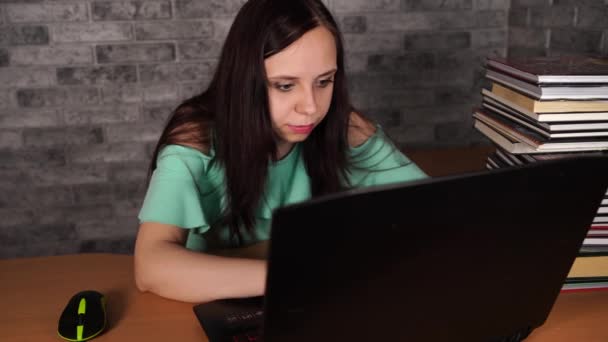 Молодая женщина сидит за столом и пользуется ноутбуком. Концентрированная брюнетка смотрит на дисплей ноутбука во время работы на фоне серой стены — стоковое видео