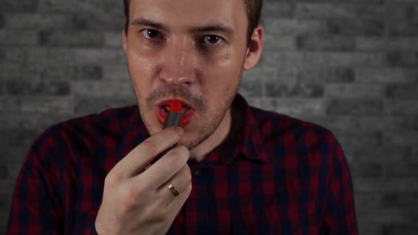 Un hombre se pinta los labios con lápiz labial rojo, contra una pared de ladrillo — Vídeo de stock