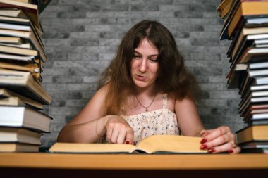 Yorgun kız öğrenci kitaplar arasında okuma. Kitap yığını ile masada oturan ve gri duvarın arka planda okuma Pensive genç kadın