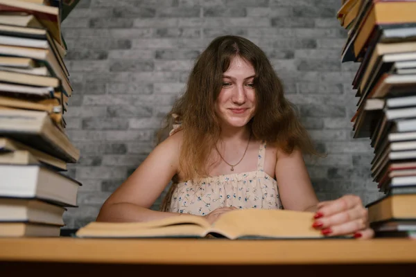 Втомлена студентка читає серед книг. Молода жінка сидить за столом з купою книг і читає на тлі сірої стіни — стокове фото