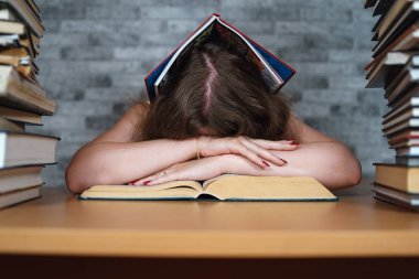 Yorgun kız öğrenci kitaplar arasında okuma. Kitap yığını ile masada oturan ve gri duvarın arka planda okuma Pensive genç kadın