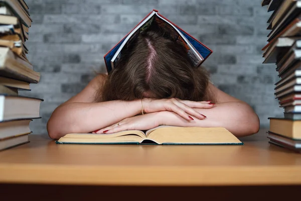 Vermoeide vrouwelijke student lezing tussen boeken. Pensive jonge vrouw zittend aan tafel met stapel boek en lezing op achtergrond van grijze muur — Stockfoto