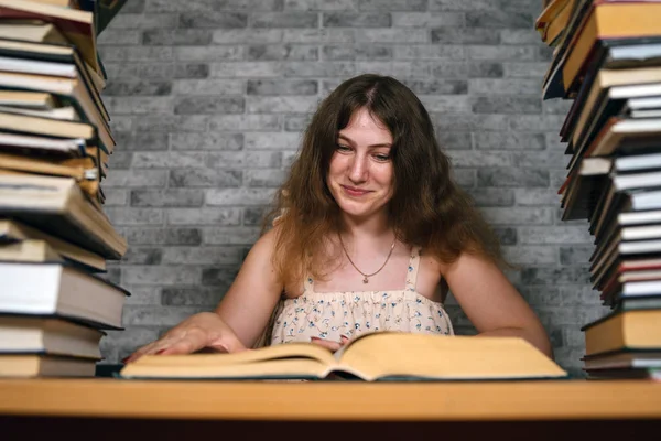 Втомлена студентка читає серед книг. Молода жінка сидить за столом з купою книг і читає на тлі сірої стіни — стокове фото