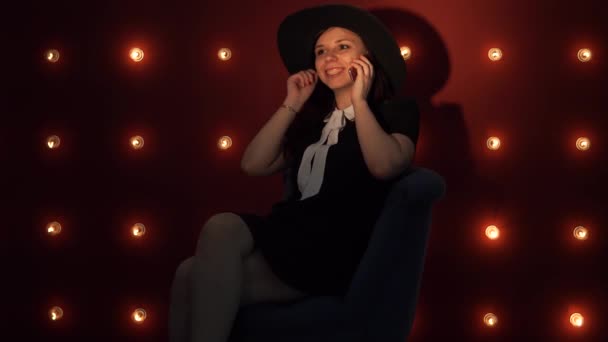 Jovem mulher de chapéu contra parede iluminada, falando usando um smartphone — Vídeo de Stock