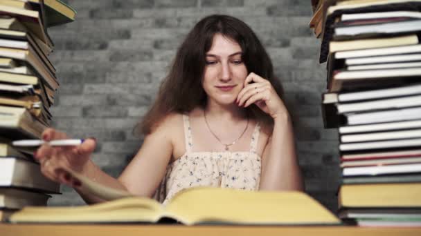 Estudiante cansada leyendo entre libros. Mujer joven pensativa sentada a la mesa con pila de libro y lectura sobre fondo de pared gris — Vídeos de Stock