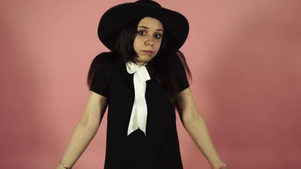 Young female in black dress and hat on a pink background. Attractive woman wearing black outfit standing on pink background in perplexity on a pink background. The confused woman shrugs — Stock Video