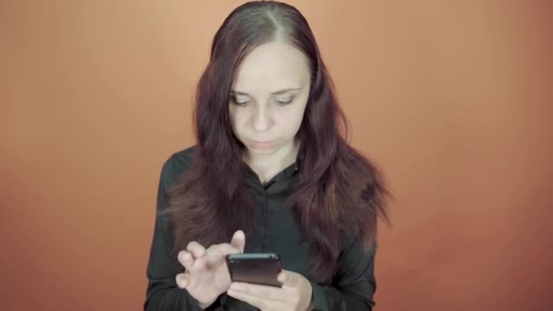 Молодая женщина держит в руке мобильный телефон на оранжевом фоне. Женщина печатает сообщение на смартфоне — стоковое видео