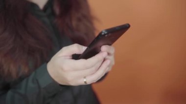 Turuncu arka planda cep telefonu tutan genç bir kadın. Akıllı telefondan mesaj yazan bir kadın.