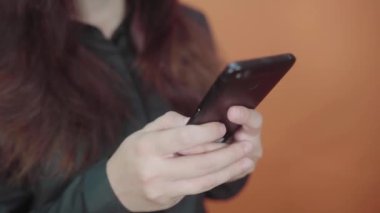 Turuncu arka planda cep telefonu tutan genç bir kadın. Akıllı telefondan mesaj yazan bir kadın.