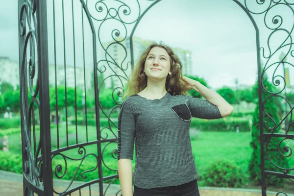 Улыбающаяся женщина против забора в саду. Содержание женщина смотрит мечтательно далеко, стоя в зеленом парке против декоративного железного забора — стоковое фото
