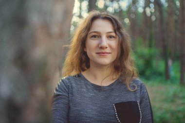 Ağacın arkasından dışarı bakan ciddi bir kadın. Sakin bir kadın ağaca dokunuyor ve sessiz ormanda kameraya bakıyor.