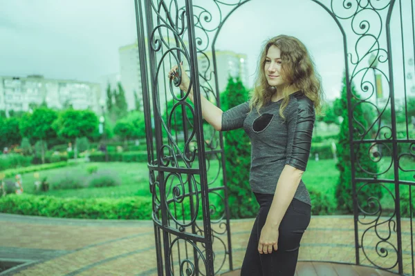 Mulher sorridente contra vedação no jardim. Conteúdo Mulher olhando sonhadoramente enquanto está em pé no parque verde contra cerca de ferro ornamental — Fotografia de Stock