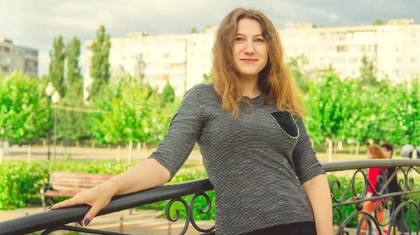 Mujer contenta mirando soñadoramente de pie en el parque verde contra la cerca de hierro ornamental. Mujer tímida apoyada en asidero en el jardín — Foto de Stock