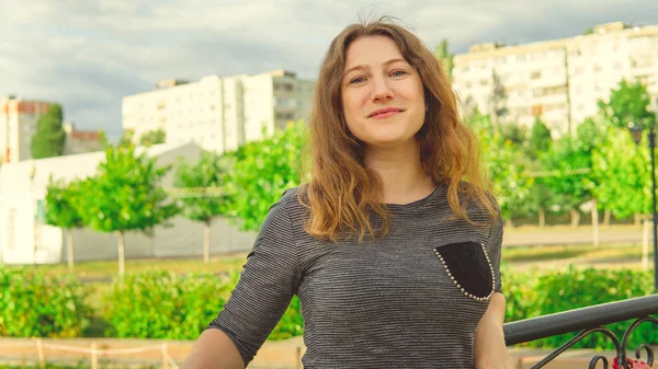 Mujer contenta mirando soñadoramente de pie en el parque verde contra la cerca de hierro ornamental. Mujer tímida apoyada en asidero en el jardín — Foto de Stock