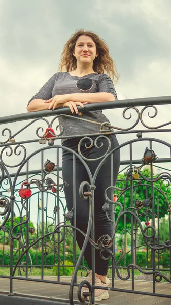Mulher conteúdo olhando sonhadoramente de pé no parque verde contra cerca de ferro ornamental. Mulher tímida apoiando-se no handhold no jardim — Fotografia de Stock