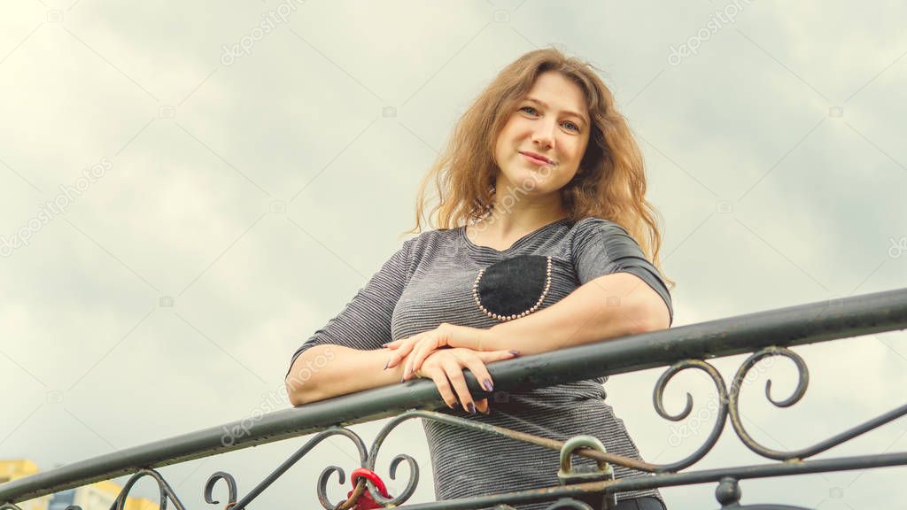 Content woman looking dreamily standing in green park against ornamental iron fence. Shy woman leaning on handhold in garden