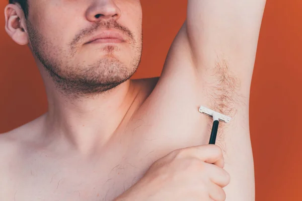 Attraktiver Mann mit blauem Rasiermesser in den Händen rasiert sich die Haare mit vereinzelten Achseln auf orangefarbenem Hintergrund — Stockfoto