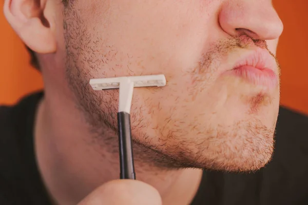 Attraktiver Mann mit blauem Rasiermesser in den Händen rasiert sich die Haare mit vereinzelten Achseln auf orangefarbenem Hintergrund — Stockfoto