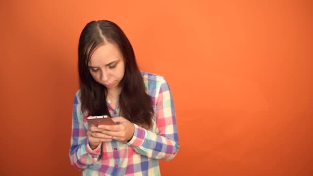 ผู้หญิงสาวถือโทรศัพท์มือถือในมือบนพื้นหลังสีส้ม ผู้หญิงกําลังพิมพ์ข้อความบนสมาร์ทโฟน — วีดีโอสต็อก